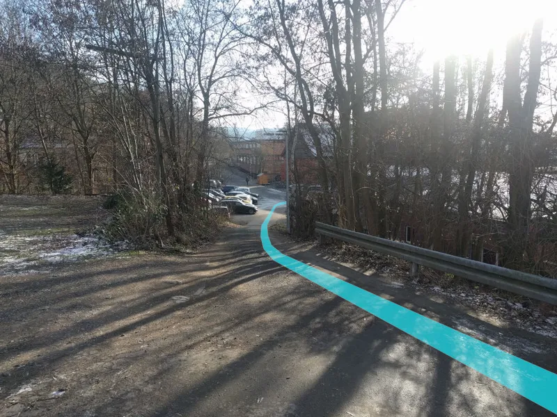 Ein unbefestigter Weg an einer Leitplanke; ein türkiser Streifen führt in einer Kurve bergab zu einem Parkplatz.