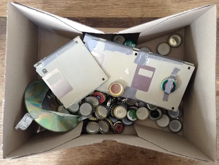 Blick von oben in einen Pappkarton mit einem Haufen Kronkorken, drei mit Klebeband zusammengeklebten 3,5-Zoll-Disketten und einer zerbrochenen CD.